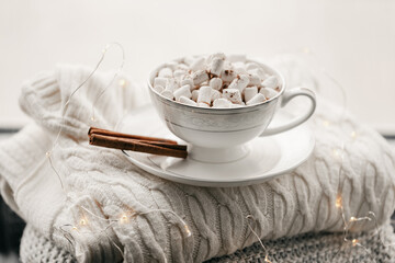 Still life details, cup of coffee in living room, top view point. Winter weekend with a book on the sofa. White knitted sweaters are stacked. Delicate marshmallow with cinnamon in a mug with coffee