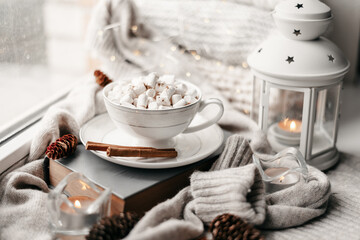 Still life details, cup of coffee in living room, top view point. Winter weekend with a book on the sofa. White knitted sweaters are stacked. Delicate marshmallow with cinnamon in a mug with coffee