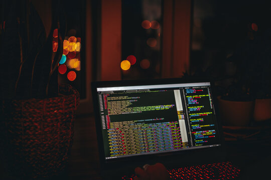A young man programmer coding on a laptop in the dark with a view of the lights of the night city, color lighting in the room, home decor