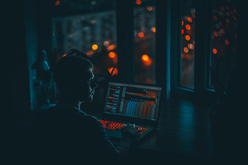 A young man programmer coding on a laptop in the dark with a view of the lights of the night city,...