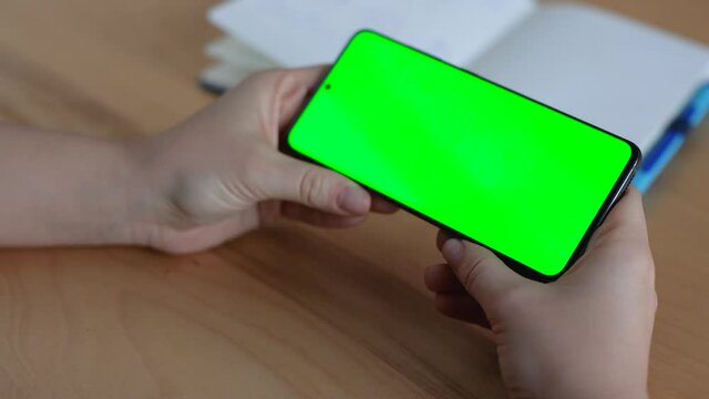 Female`s Hands are holding a Smartphone with Green Screen on the Desk. Concept of Studying, Working Indoor, Online. Woman using Smartphone, watching Chroma Key online, reading Social Media at home.