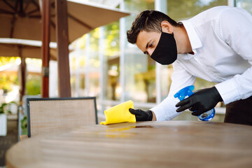 Waiter in protective face mask and gloves cleaning the table with disinfectant spray in a restaurant. Covid- 2019.