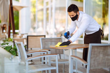 Waiter in protective face mask and gloves cleaning the table with disinfectant spray in a restaurant. Covid- 2019.