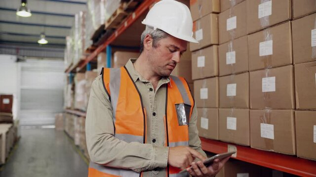 Caucasian worker typing on digital tablet assorting parcels according to checklist standing in factory warehouse 