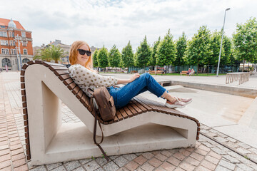 Beautiful young woman resting on a bench at the city background. Girl relax in a sunny day. Travel and active life concept.