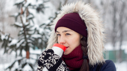 Beautiful young girl enjoys drinking coffee from a thermos while walking in a winter park.