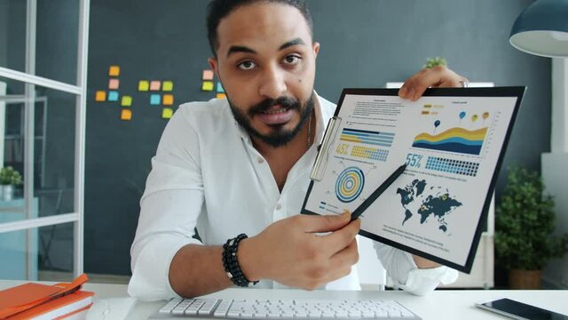 Portrait of mixed race guy talking and showing charts during video conference online in office. Modern communication and business people concept.