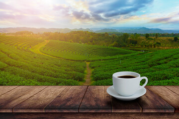 White cup of hot coffee on wooden table with tea plantation with sun light background