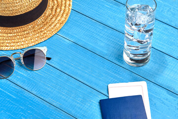 Fototapeta na wymiar Straw hat, sunglasses, glass of water with ice cubes and passport with ticket on blue wooden background. Travelling and tourism. Close up, copy space