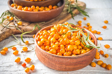 Sea buckthorn berries in small organic wooden bowl, with sea buckthorn branches on white wooden desk background - obrazy, fototapety, plakaty