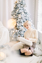 Happy positive Caucasian blond woman in trendy sweater and skirt, sitting indoors on the floor with cozy Christmas decorations, pillows, opening a Christmas box. Christmas tree on the background