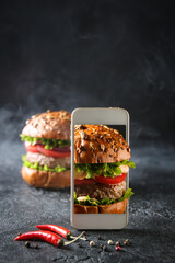 American cuisine. Burger with meat, lettuce in your mobile phone. Mobile photo of fast food on a black background with smoke. Chili peppers and spices. Background image, copy space
