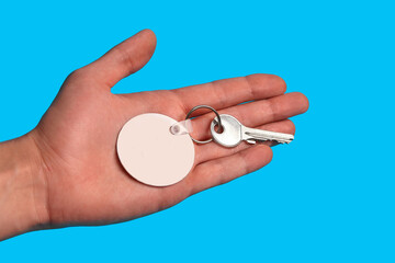 Key with blank white plastic keychain on metal ring on palm of unknown man posing against blue...