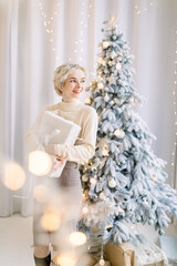 Portrait of smiling Caucasian joyful woman, wearing sweater and skirt, and holding white present box, standing near decorated Christmas tree and looking towards