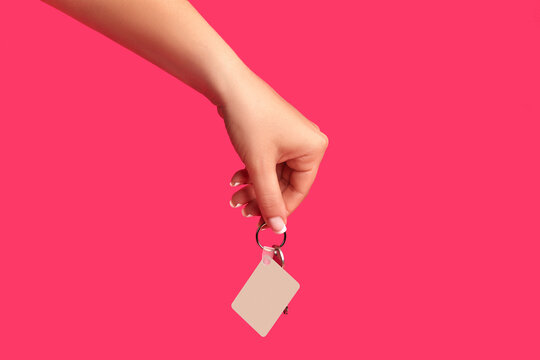 Hand of unrecognizable female is holding a key with empty white square plastic key fob on metal ring against pink background. Close up, copy space