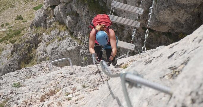 Woman with red backpack and helmet climbs vertical section on a via ferrata route in Romania, above a suspended bridge. Adventure, active people, top down view.