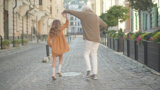 Wide shot back view of joyful little Caucasian girl jumping as walking with senior grey-haired man on city street. Cheerful grandfather and granddaughter having fun on weekends outdoors.