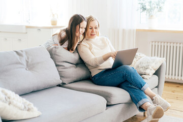 Mom and daughter spend time at home on the couch in the living room with a computer. Online communication and education.