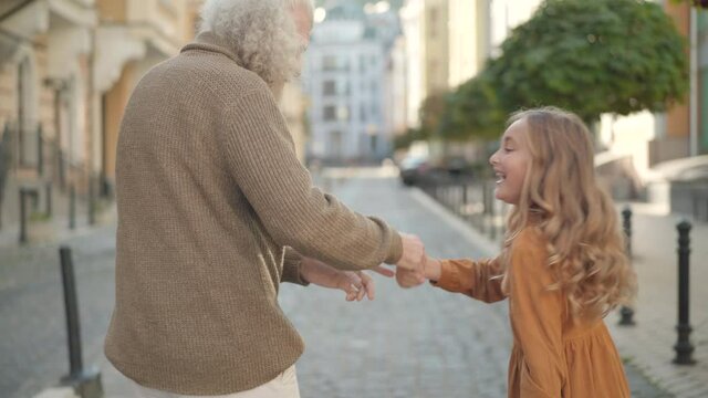 Back view of carefree Caucasian grandfather and granddaughter strolling and dancing on city street. Happy cute girl hugging adorable grandparent and smiling. Happiness and childhood.