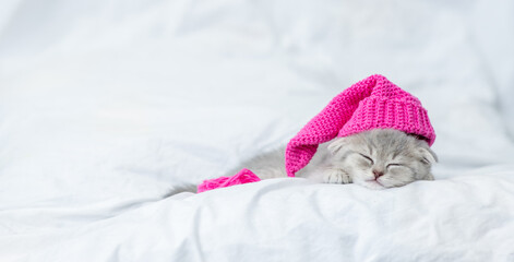 Fototapeta na wymiar Cute baby kitten wearing warm hat sleeps on a bed at home. Empty space for text
