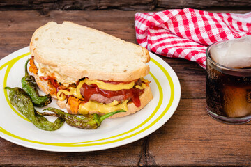 Hamburguesa con ketchup y mostaza entre dos rebanadas de pan, pimientos fritos en el plato, un vaso con bebida de cola en la mesa, fondo rustico
