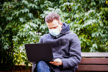 Handsome young European man in a park with laptop with a medical face mask on. Freelance working outside the office during an Covid-19 epidemic. Selective focus