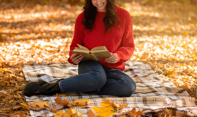 Unrecognizable millennial woman in casual fall clothes sitting on picnic blanket and reading captivating book outdoors