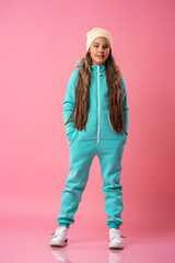 Teen girl in a beanie, warm jumpsuit with sneakers stands on a pink background