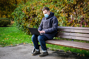 Handsome young European man in a park with laptop with a medical face mask on. Freelance working outside the office during an Covid-19 epidemic. Selective focus