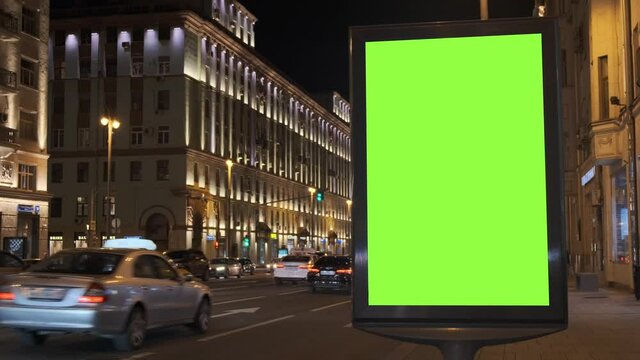A green screen billboard located on the sidewalk on which people walk, next to a busy street. Cars move in the evening. 4K