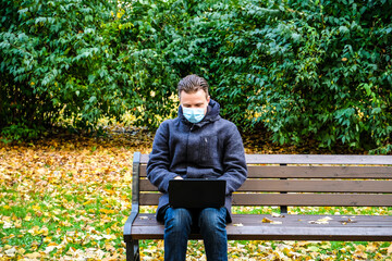 Obraz premium Handsome young European man in a park with laptop with a medical face mask on. Freelance working outside the office during an Covid-19 epidemic. Selective focus