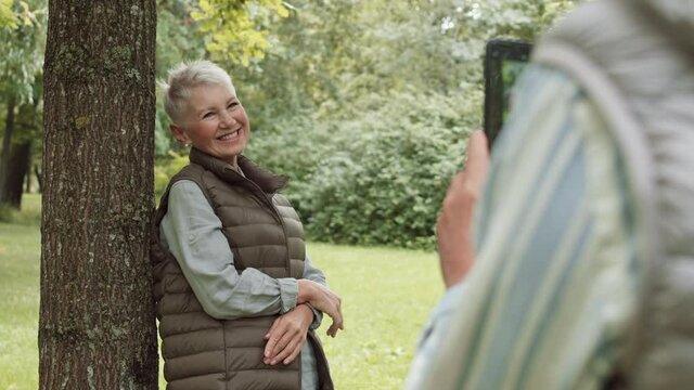 Over shoulder of unrecognizable man holding smartphone taking pictures of his senior white-haired Caucasian wife. Woman standing by tree trunk in park, posing and smiling on camera