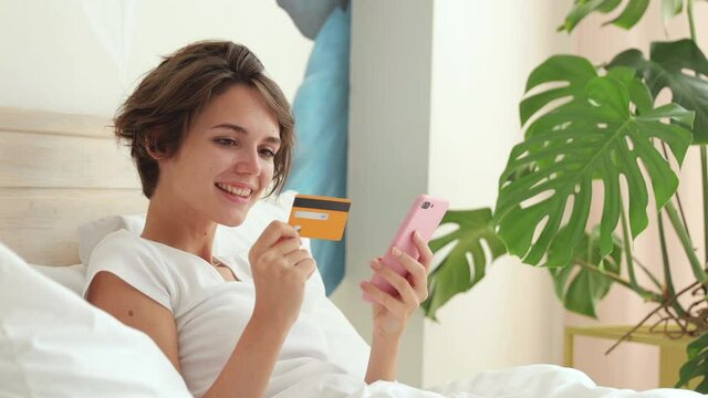 Young woman in white t-shirt pajamas home wear lying in bed spending time in bedroom hold in hand credit bank card using mobile cell phone makes purchases online shopping. Rest relax lifestyle concept