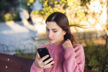 Young woman in pink sweater looking at her smartphone and sitting on a bench