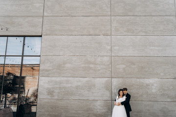 Newlyweds hugging on a gray wall background. Groom is embracing bride from the back. The bride is dressed in a classic dress and veil. Groom with beard dressed in a classic black suit. Copy space