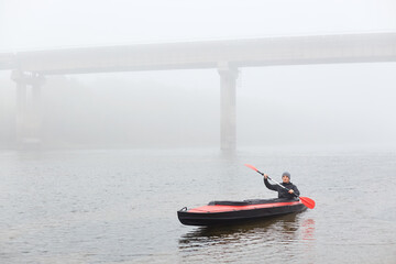 Man canoeing in traditional wooden kayak on large lake at cold cloudy day, fog over water, handsome guy wearing black jacket and gray cap, kayaking alone in lake with bridge on backround.