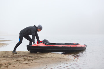 Man pulling canoe into water, wearing black clothing, posing on bank of river, handsome sports man doing water sport in foggy autumn day, canoeing in cloudy morning.