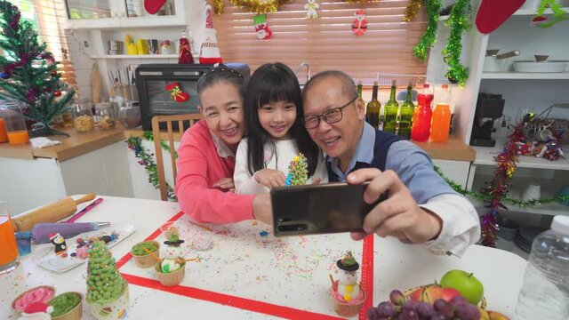 Happy Asian grandparents and cute little granddaughter taking a selfie together with Christmas tree shaped cupcakes which they've just made together in Christmas decorated kitchen at home.