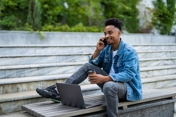 Obraz premium Young african american man sitting on the bench with laptop and talking on the phone
