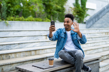Smiling young african man in casual clothes making video call with smartphone and gesturing greeting while sitting on bench in park