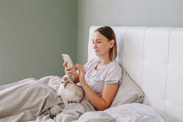 Beautiful middle age woman sitting in bed in morning with a smartphone surfing Internet. Young woman talking with friends in social media. Work from home. Digital online Internet addiction. - 388568065