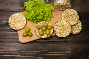 On a dark wooden background there is a cutting board, pieces of round whole grain bread, olives, lettuce.