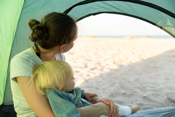 Mom with little baby sitting in the tourist tent. Sea and beach views from the camping tent