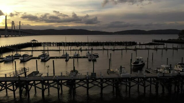 A low angle shot of the Tarrytown Marina in upstate NY. The Mario M. Cuomo Bridge in in the background as the drone truck left & boom up. The setting sun shines on the Hudson River.