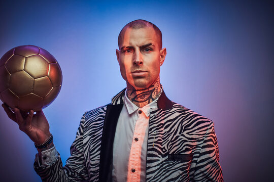 Stylish and succesful football trainer dressed in custom suit with jewellery poses with golden ball in abstract light background.