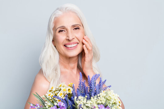 Photo of beautiful retired woman hold big bunch wild flowers touch cheek floral cream isolated grey color background
