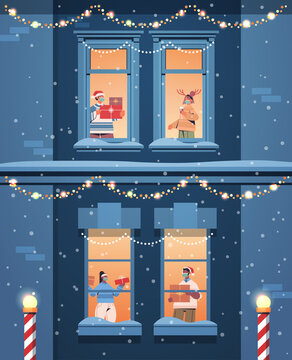 People In Santa Hats Holding Gifts Mix Race Neighbors Standing In Window Frames New Year Christmas Holidays Celebration Self Isolation Concept Building House Facade Vertical Vector Illustration