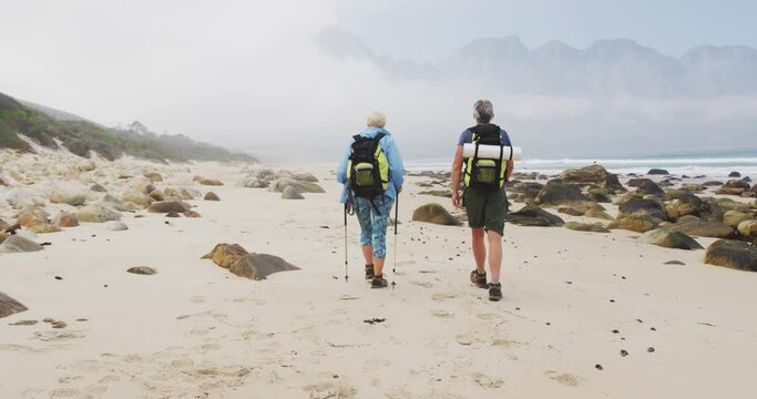 Rear view of senior hiker couple with backpack and hiking poles walking while hiking on the beach