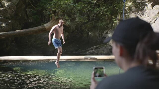 Man jumps off of log into swimming hole, woman takes photo on phone