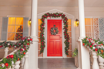 Fototapeta premium Christmas porch decoration idea. House entrance with red door decorated for holidays. Red and green wreath garland of fir tree branches and lights on railing. Christmas eve at home
